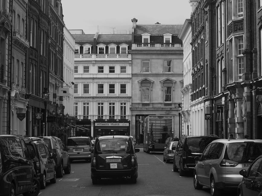 Black and white photograph of a narrow urban street, likely in London, with parked vehicles lining both sides of the road. In the foreground, a small black van is parked parallel to the curb, with oth