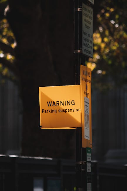 Close-up image of a metal parking restriction signpost mounted on a concrete wall beneath a row of windows at night. The top sign indicates no parking except with a permit from 7 am to 6 pm, Monday to