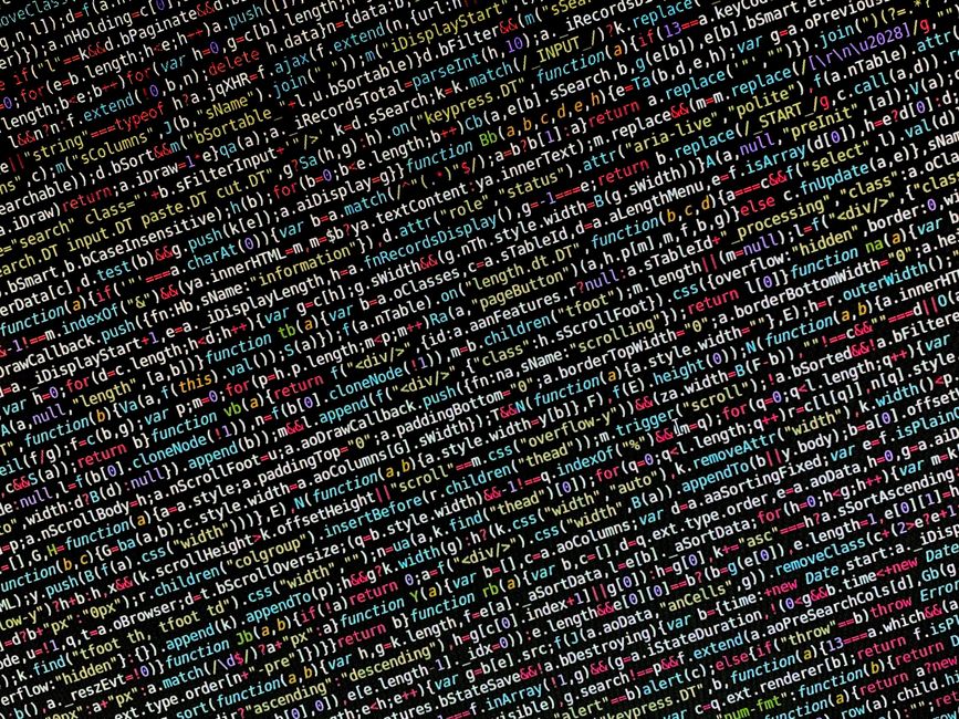 A black laptop computer with a glowing red and blue keyboard is positioned on a desk, displaying lines of code or data on its screen, which is slightly out of focus. Next to the laptop, there is a whi