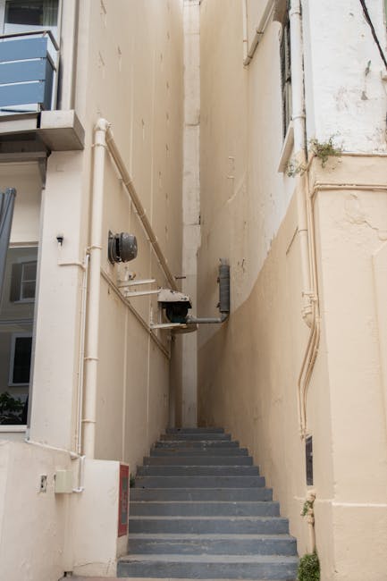 A narrow staircase with dark grey steps ascending between two tall, plain cream-colored walls, located outside a building in an urban setting. On the left wall, there are visible white pipes running v