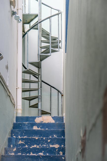 A narrow staircase with dark grey steps ascending between two tall, plain cream-colored walls, located outside a building in an urban setting. On the left wall, there are visible white pipes running v